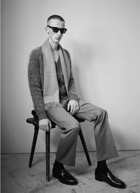 A man in sunglasses sitting on a stool wearing a cardigan, scarf, and dress pants is featured in this grey-scale studio fashion shot.