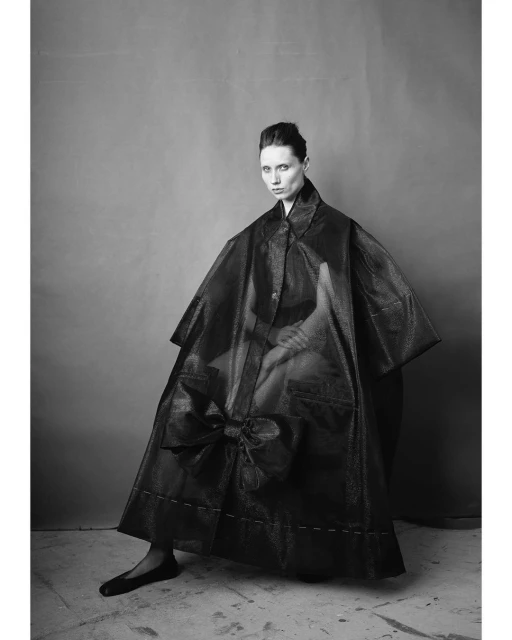 Monochrome portrait of a woman in a cape dress with a bow, styled in a photo studio with grey backdrop, emphasizing fashion and portraiture.