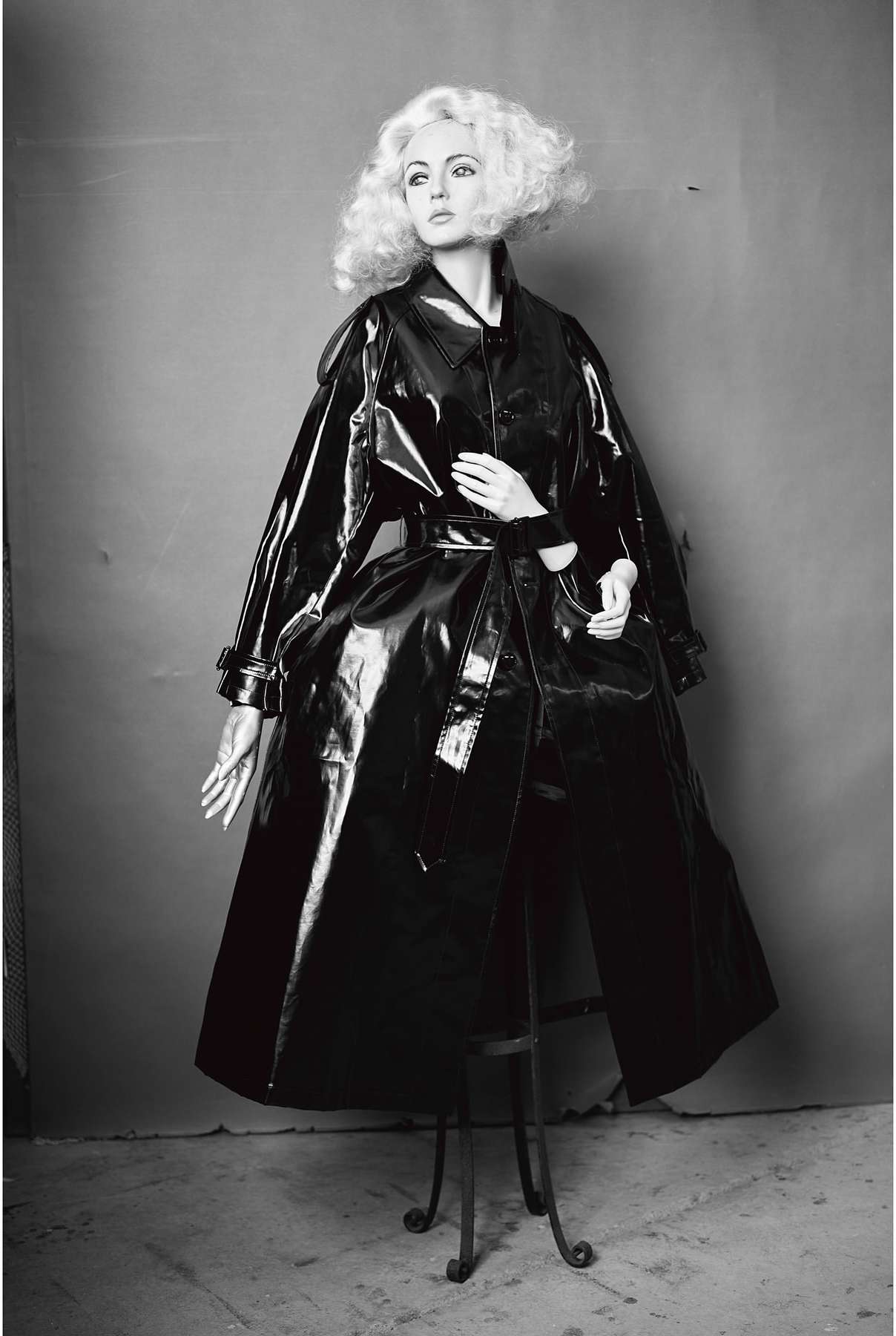 Monochromatic shot of a fashion model with a blonde wig wearing a glossy black vinyl trench coat on a metal stool against a grey backdrop.