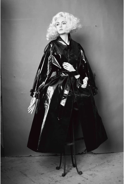 Monochromatic shot of a fashion model with a blonde wig wearing a glossy black vinyl trench coat on a metal stool against a grey backdrop.