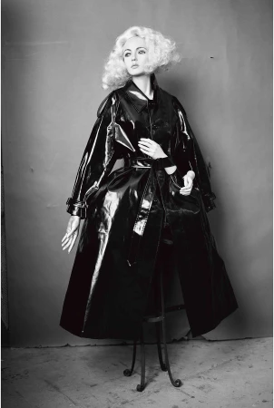 Monochromatic shot of a fashion model with a blonde wig wearing a glossy black vinyl trench coat on a metal stool against a grey backdrop.
