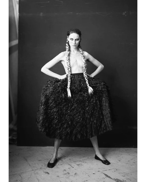 Black and white portrait of a slender woman with long braids wearing a textured black skirt and ballet flats in a photo studio.