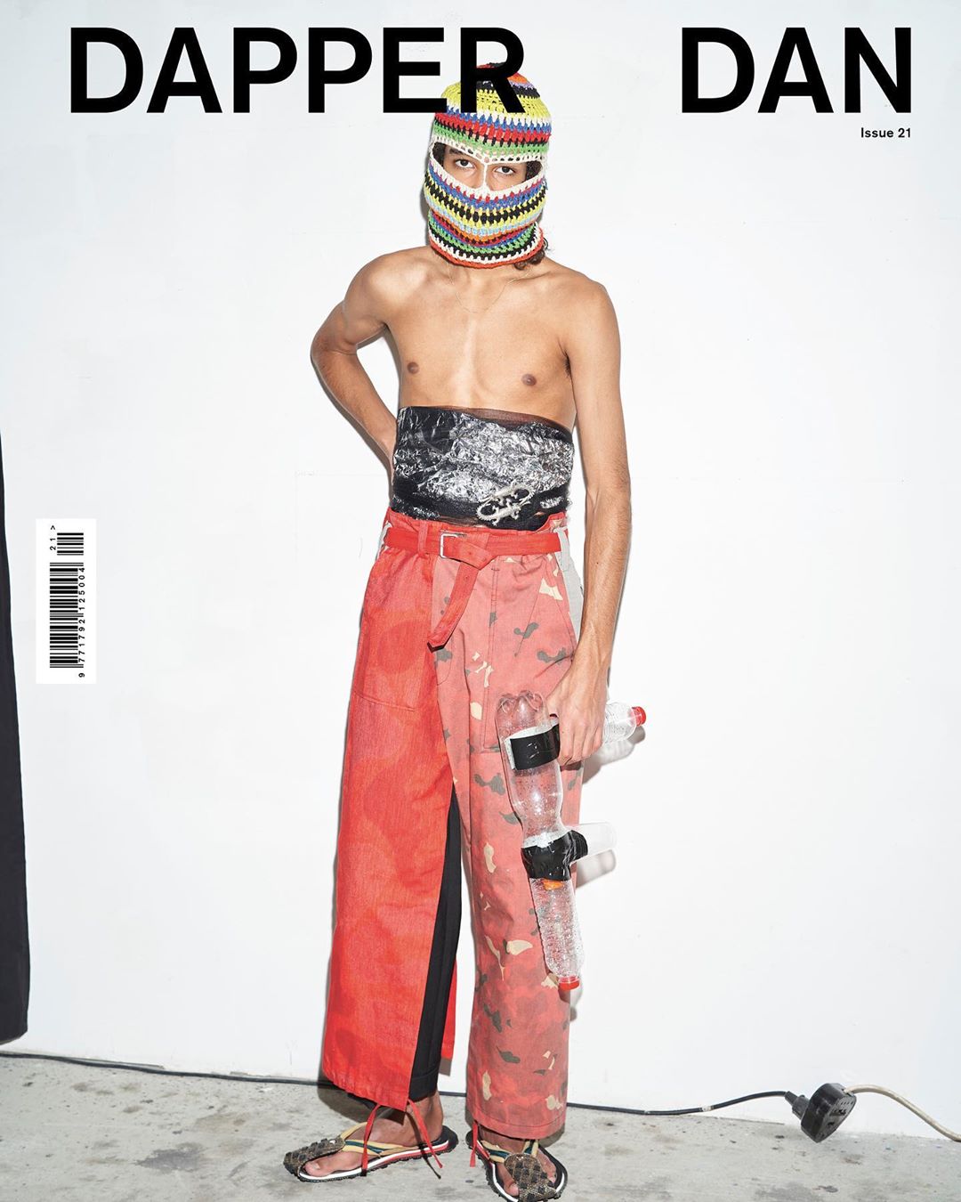 Magazine cover featuring a person with a knitted balaclava, decorative chest wrap, orange pants and water bottle accessories against a white backdrop.
