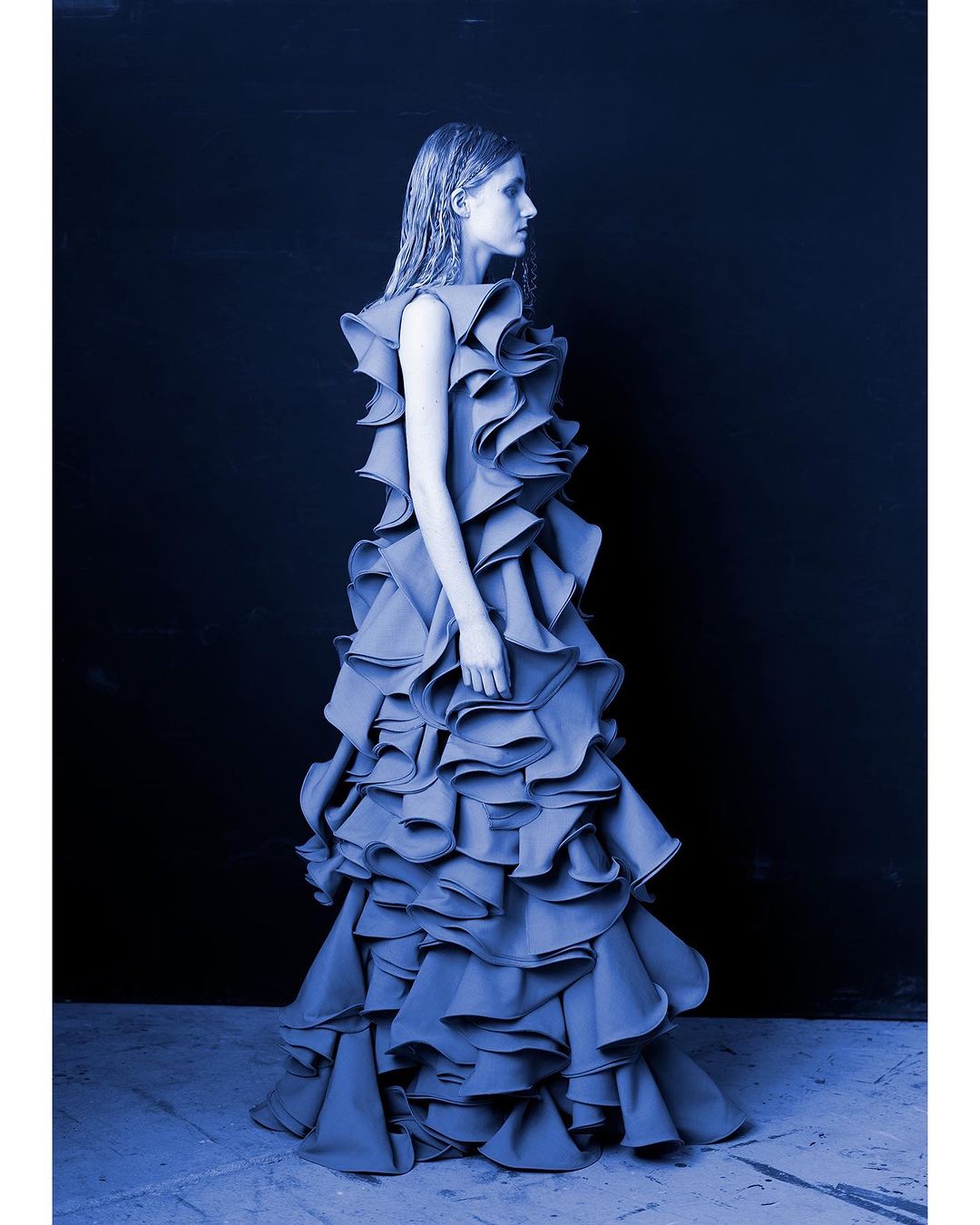 Model wearing a heavily ruffled sleeveless blue gown standing against a dark backdrop in profile