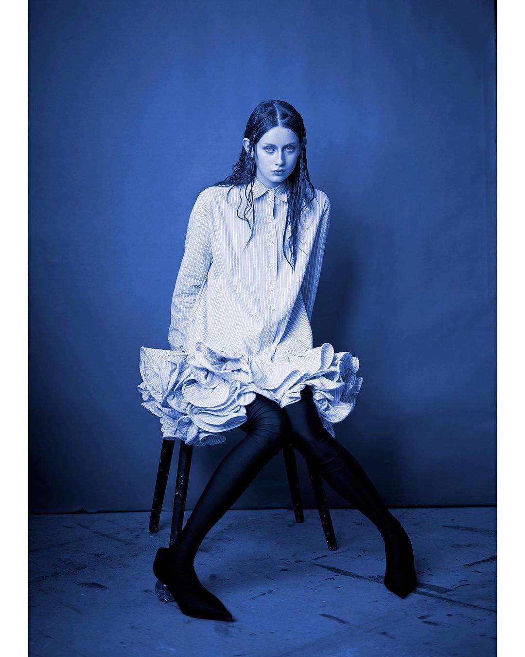 Young woman in a striped shirt dress poses on a stool against a blue background in a studio shot with dramatic lighting.
