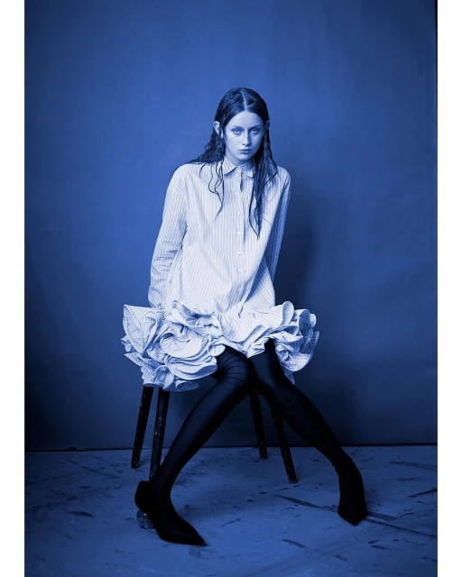 Young woman in a striped shirt dress poses on a stool against a blue background in a studio shot with dramatic lighting.