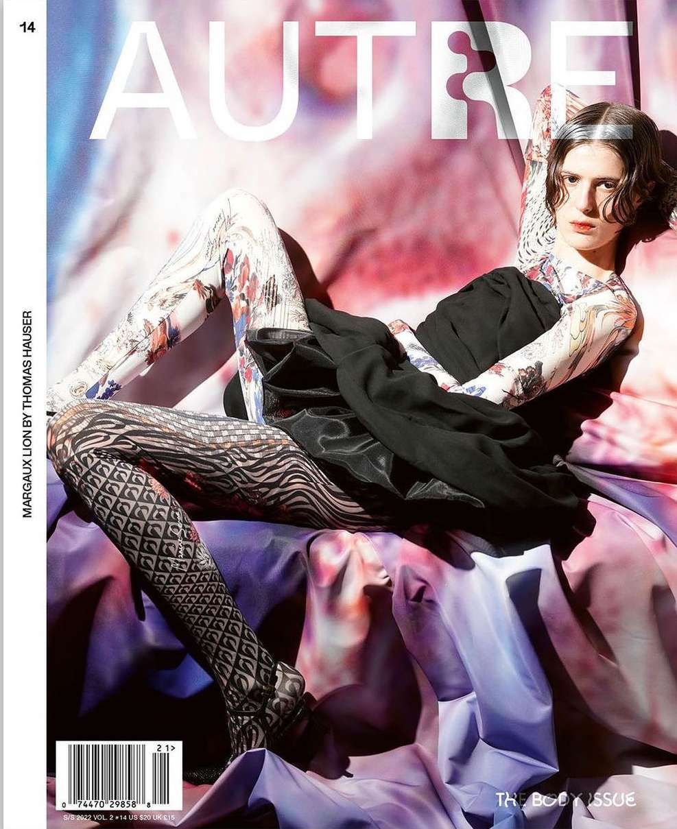 Magazine cover featuring a person with dark hair and tattoos, wearing a black dress and patterned tights, reclining against fabric.