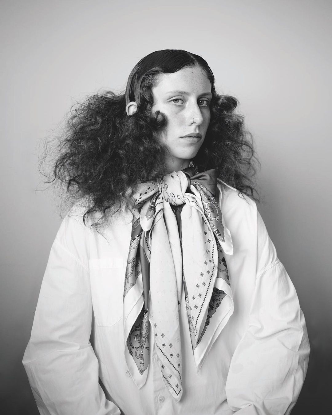 Black and white portrait of a young adult woman with curly hair and a patterned scarf wearing an oversized white shirt in studio light.