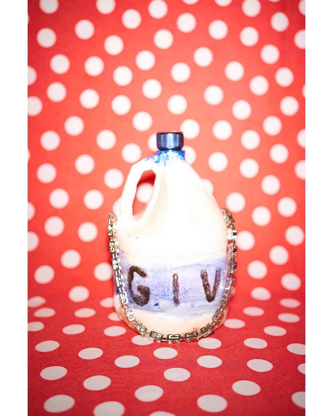 Still life of altered plastic jug adorned with costume jewelry on a vibrant red polka dot background, emphasizing altered materials.
