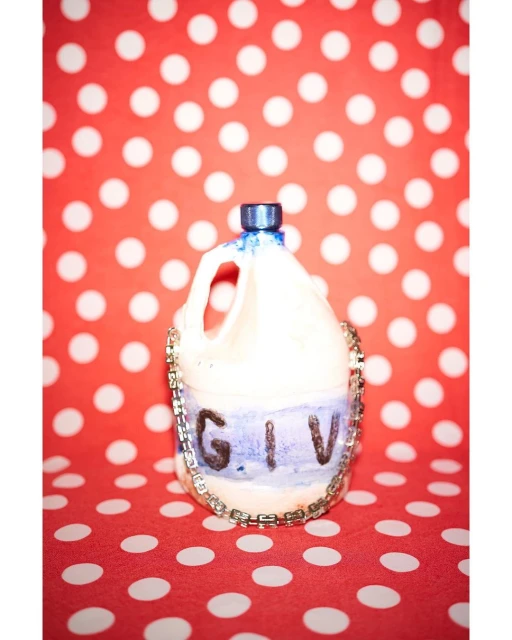 Still life of altered plastic jug adorned with costume jewelry on a vibrant red polka dot background, emphasizing altered materials.