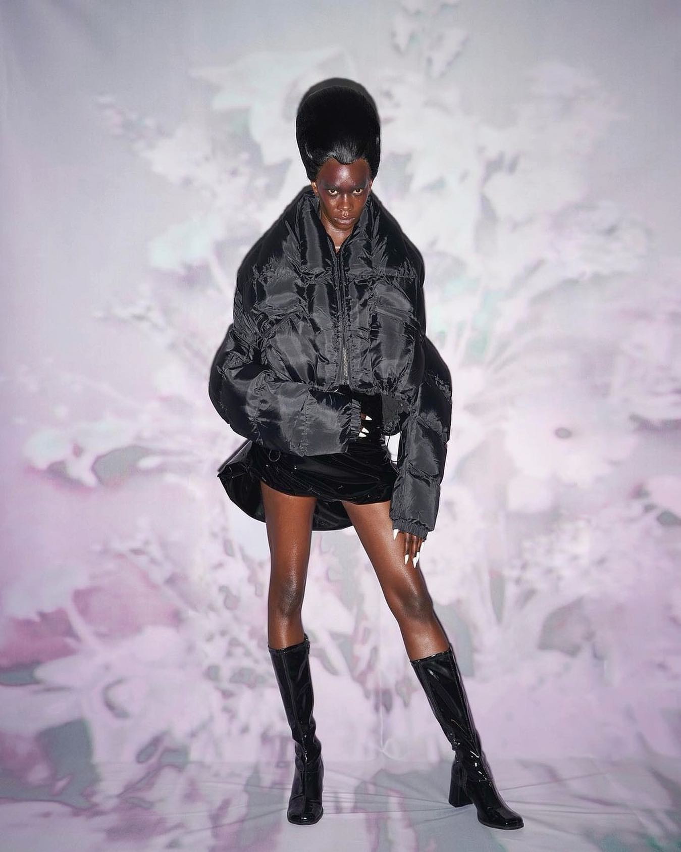A striking fashion portrait featuring a Black woman in a black puffer jacket, mini skirt and knee-high boots, set against a floral backdrop.
