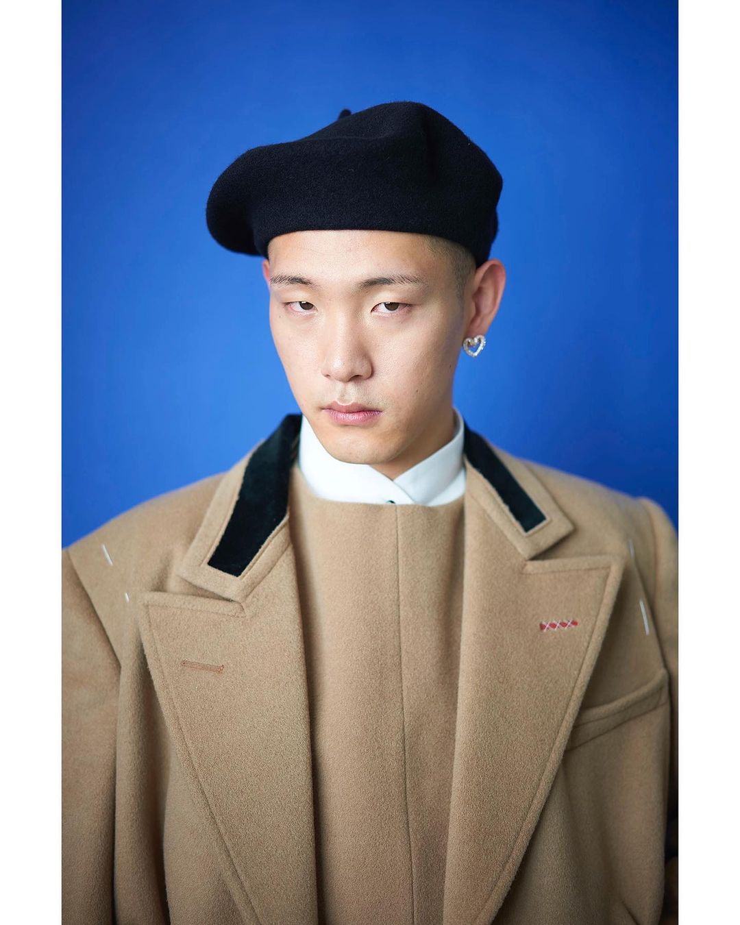 Portrait of adult male with buzz cut, tan overcoat, beret, and heart earring against a blue studio backdrop. Fashion editorial photography.