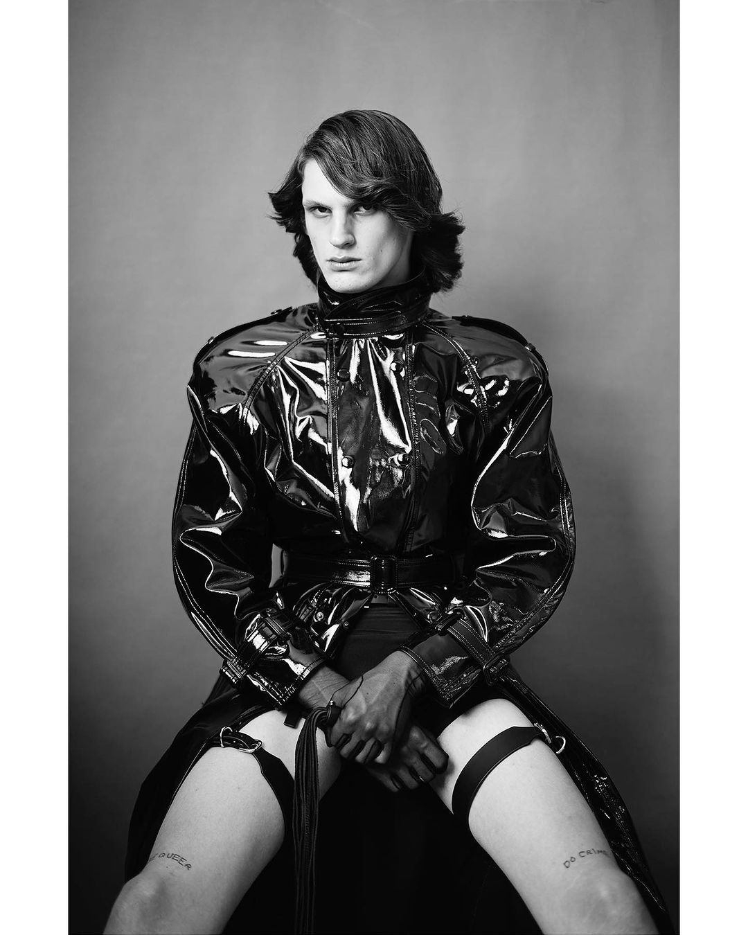 Person in a black patent leather coat, gloves, and garter belts sitting on a stool in a studio. Black and white fashion portrait.