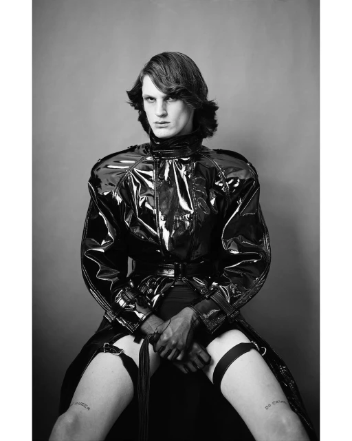 Person in a black patent leather coat, gloves, and garter belts sitting on a stool in a studio. Black and white fashion portrait.
