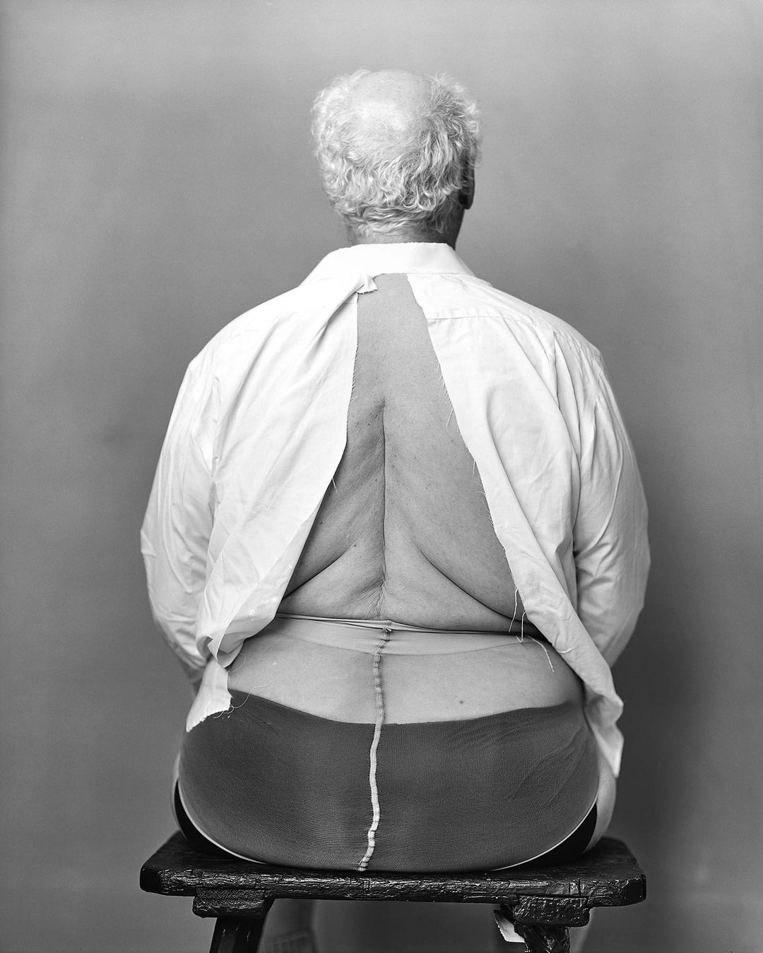 Back view of an adult, older person sitting on a stool, with a partly unbuttoned white shirt and dark tights against a gray background.