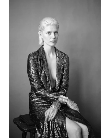 Monochrome portrait of a woman with slicked-back blonde hair wearing a sequined blazer seated on a dark stool.
