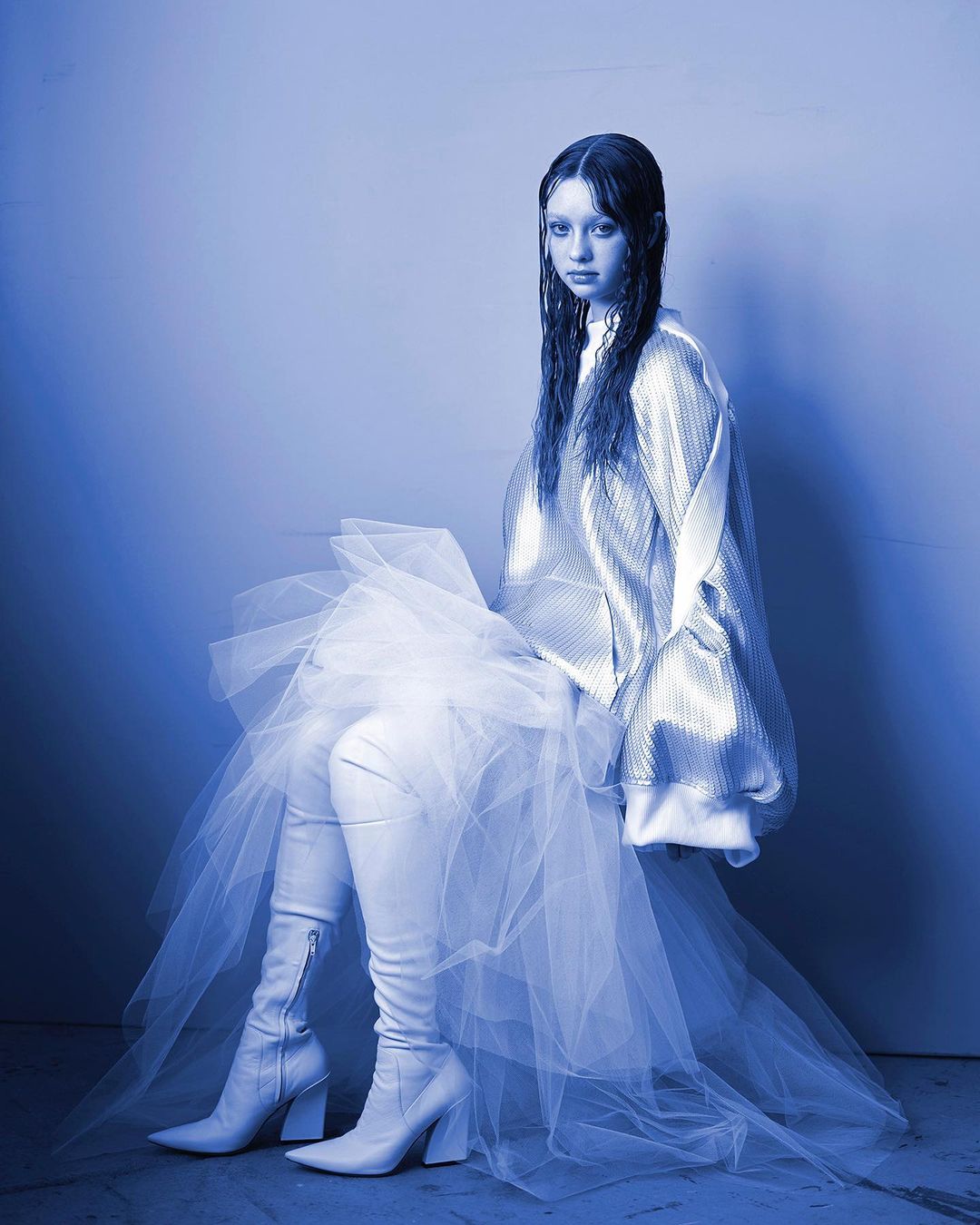 Young woman with wet hair sits in studio wearing silver jacket, long boots, and tulle skirt against gradient blue background in studio shot.