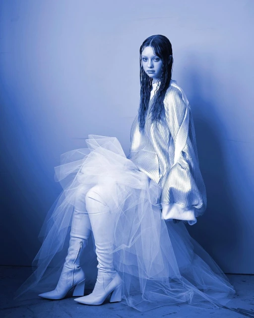 Young woman with wet hair sits in studio wearing silver jacket, long boots, and tulle skirt against gradient blue background in studio shot.