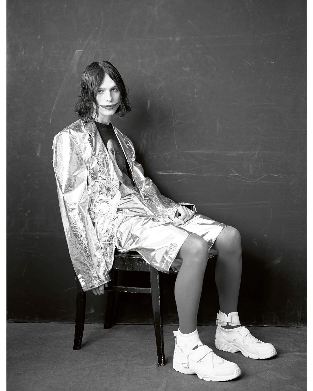 Model with dark shoulder-length hair sitting on a dark chair wearing a shiny metallic blazer shorts and white sneakers against a dark backdrop