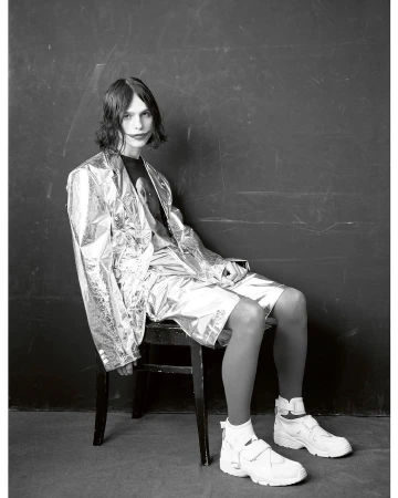 Model with dark shoulder-length hair sitting on a dark chair wearing a shiny metallic blazer shorts and white sneakers against a dark backdrop