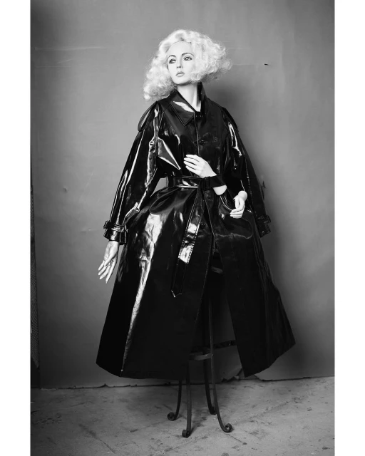 Black and white studio shot of an adult woman wearing a black patent leather trench coat and styled with short blonde curly hair.