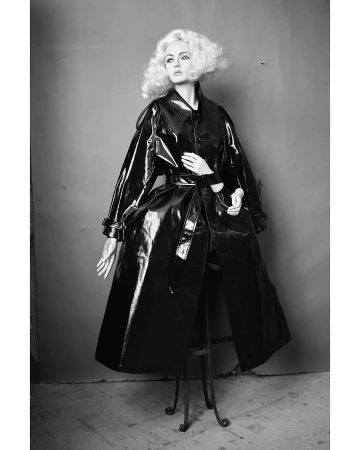 Black and white studio shot of an adult woman wearing a black patent leather trench coat and styled with short blonde curly hair.