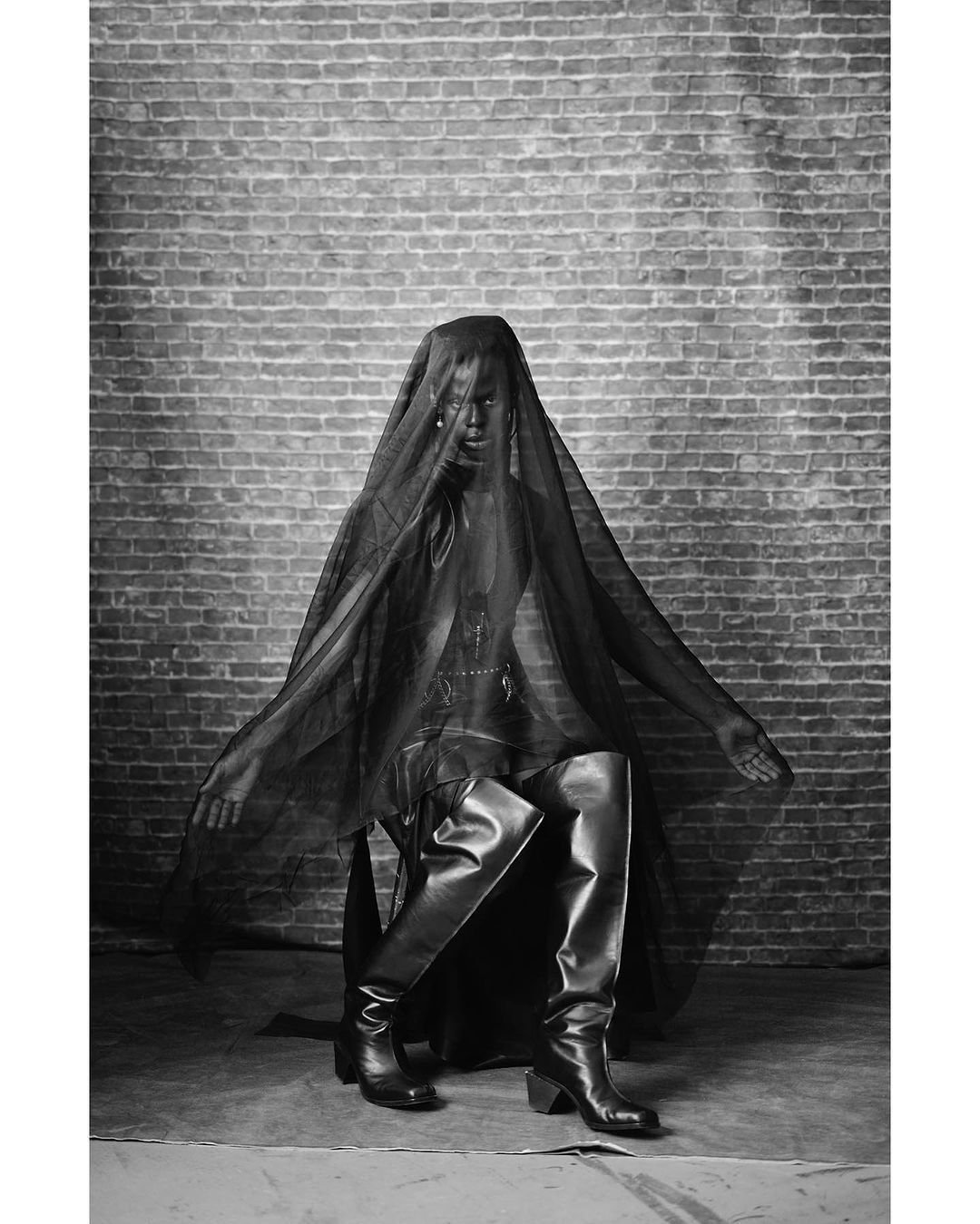 Monochrome fashion shot of a person in black clothing and thigh-high boots, seated against a brick wall and partially covered in mesh fabric.