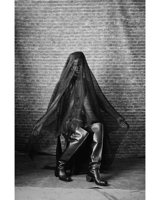 Monochrome fashion shot of a person in black clothing and thigh-high boots, seated against a brick wall and partially covered in mesh fabric.