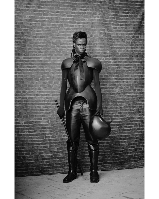Monochromatic, full-length studio shot of a singular Black woman posing in avant-garde fashion with props against a brick wall backdrop.