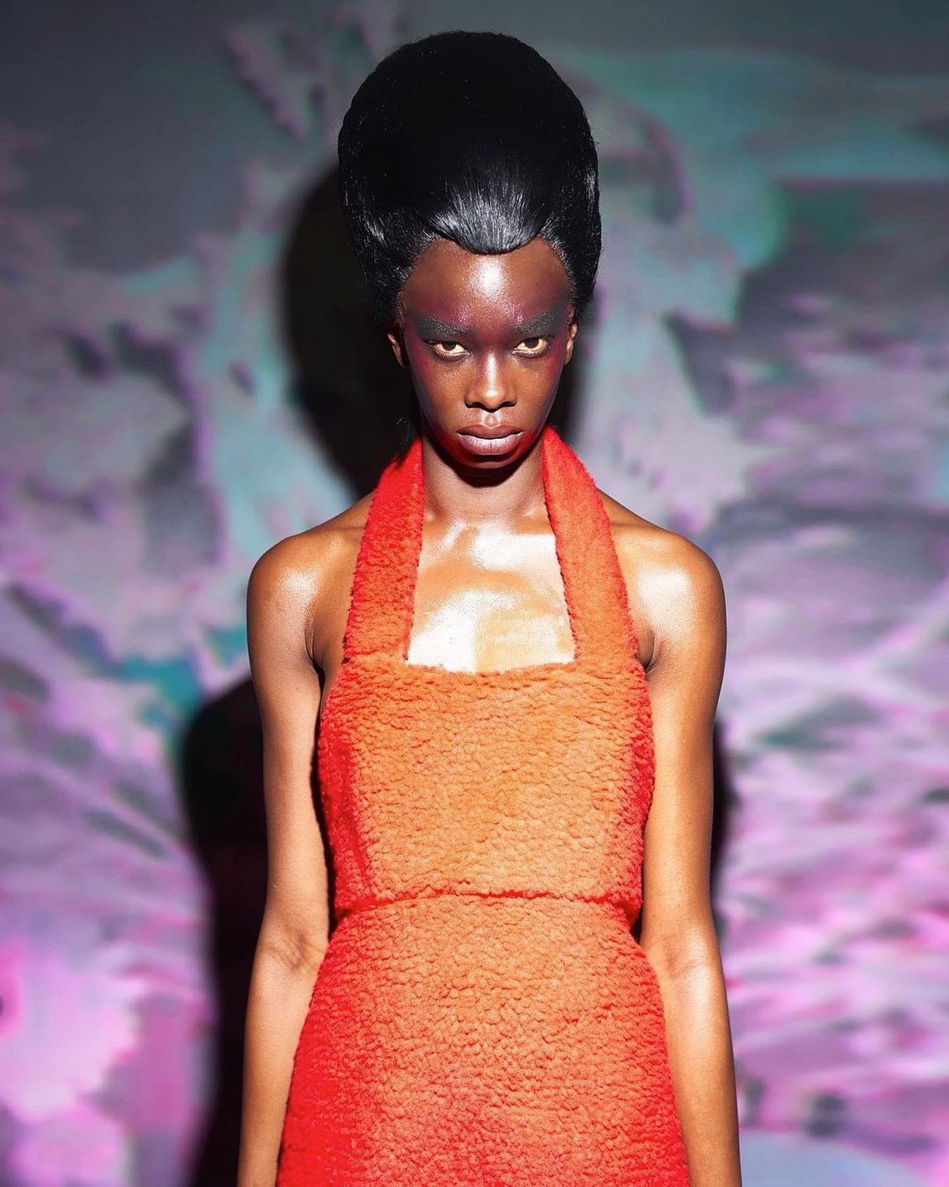 A model with dark skin and bold eye makeup poses in an orange textured dress with a surreal, colorful background.