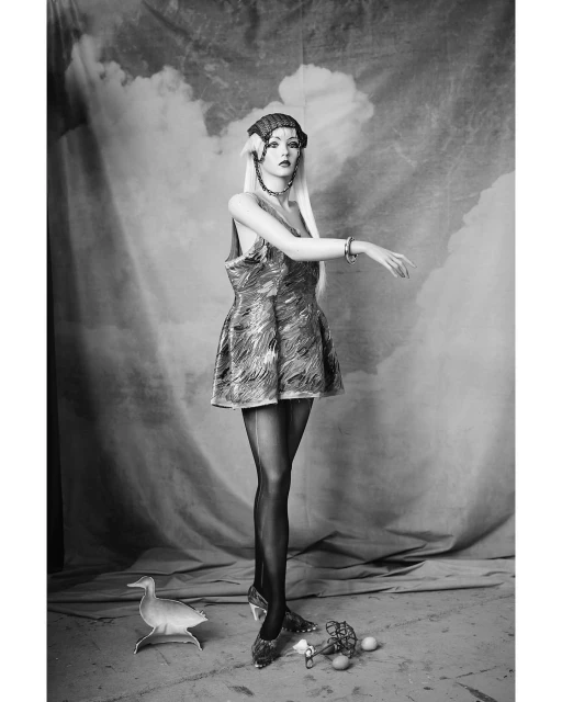 Monochrome portrait of mannequin wearing an ornate headpiece, patterned dress, sheer tights, and heels against cloudy backdrop in studio.