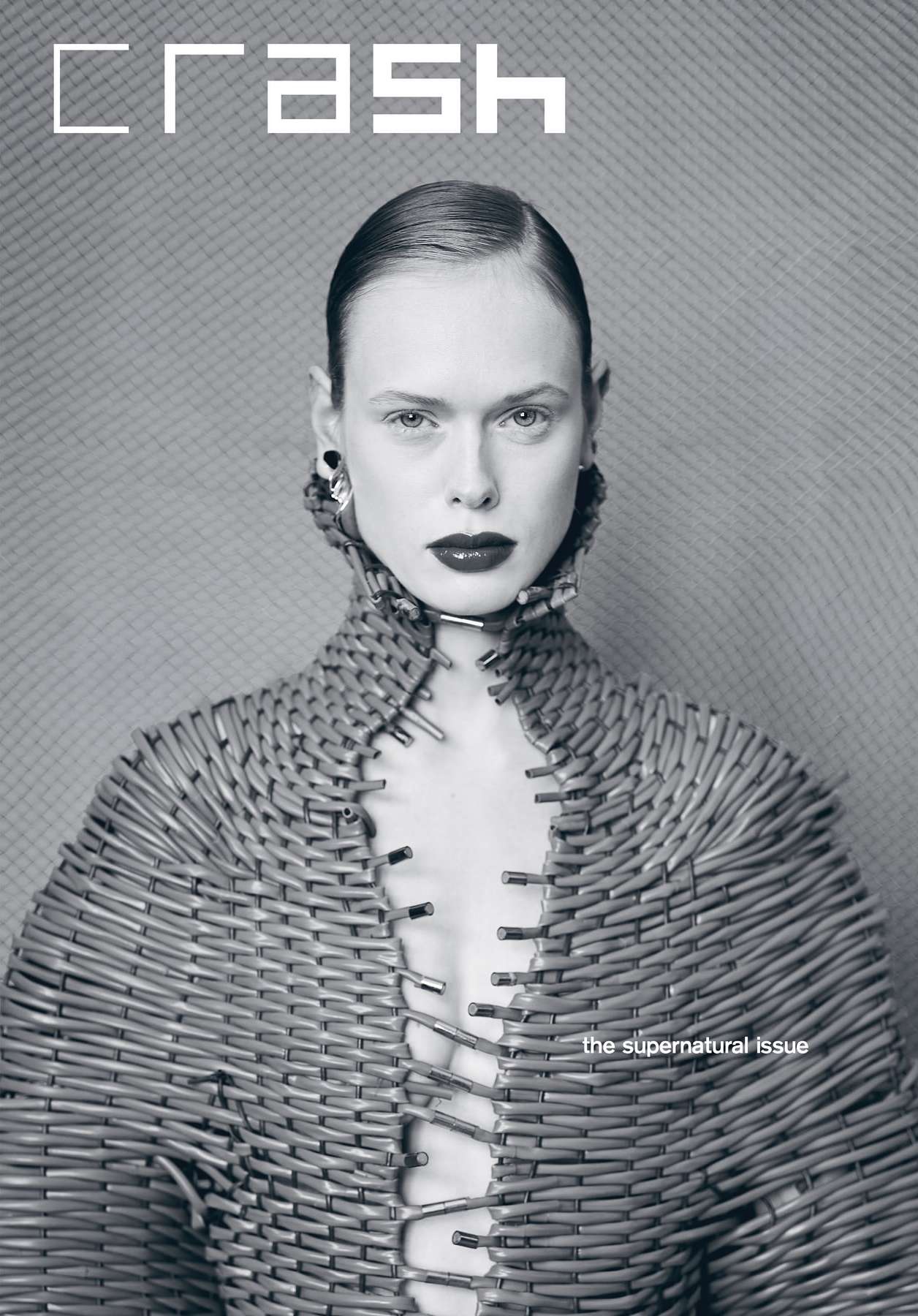 Fashion editorial image of an adult woman with slicked-back hair wearing a unique structural garment made of safety pins against a textured background.