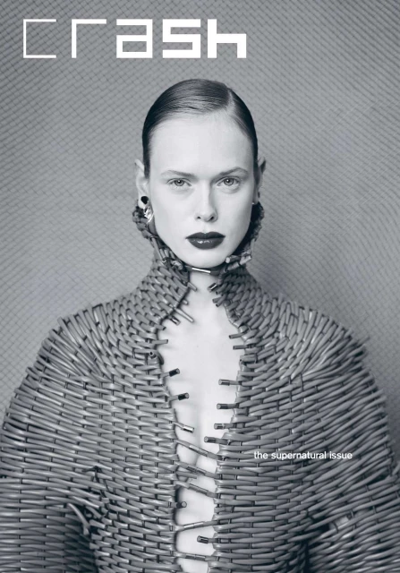 Fashion editorial image of an adult woman with slicked-back hair wearing a unique structural garment made of safety pins against a textured background.