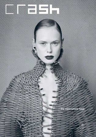 Fashion editorial image of an adult woman with slicked-back hair wearing a unique structural garment made of safety pins against a textured background.