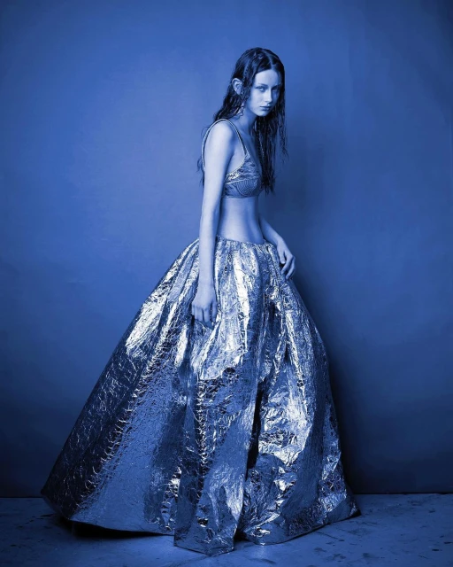 A woman in a silver bra and skirt stands in front of a blue backdrop in a studio shot. Fashion shoot with shiny material.