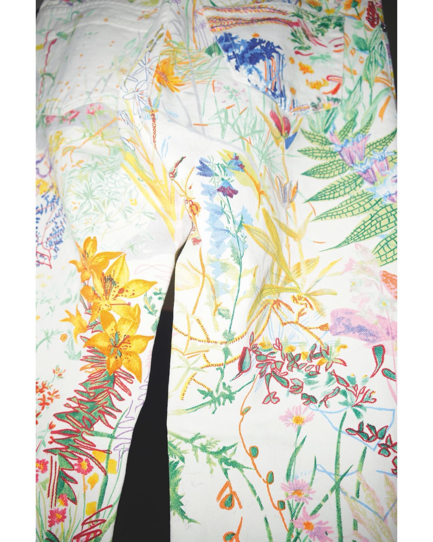White jeans with a colorful floral pattern of yellow, blue, pink, and green flowers and foliage, creating a vibrant and artistic design.