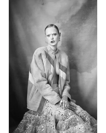 Black and white portrait of a woman wearing a patterned jacket and textured skirt against a painted studio backdrop.
