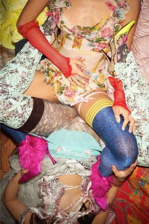 Two women are posing on a patterned surface, wearing vintage dresses, high heels with fur, lace tights, and red fishnet gloves.