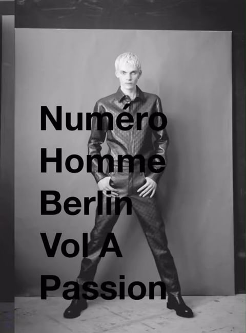 Monochrome portrait of a blonde male model standing against a grey backdrop wearing a dark leather shirt and matching trousers