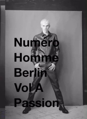 Monochrome portrait of a blonde male model standing against a grey backdrop wearing a dark leather shirt and matching trousers