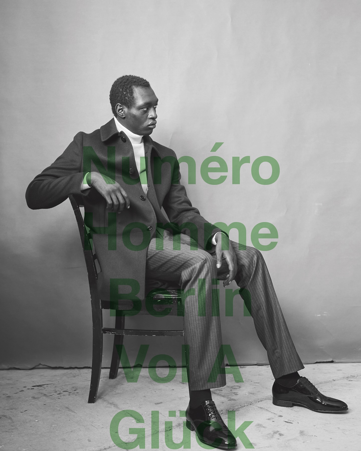 A Black male model in a suit sits in a chair against a grey backdrop, looking off to the side in a studio fashion shot.