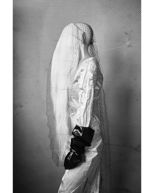 Black and white studio shot of fashion, showcasing a veiled mannequin in a white dress, accessorized with dramatic black gloves.