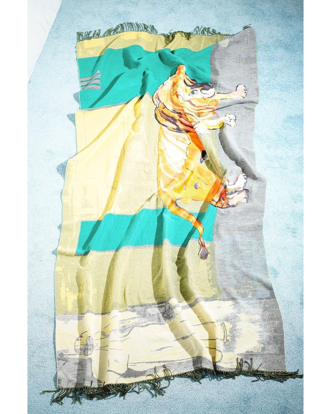 Abstract blanket with tiger graphic in yellow, orange, teal, and grey tones with fringe on both ends, flat lay on light blue backdrop.