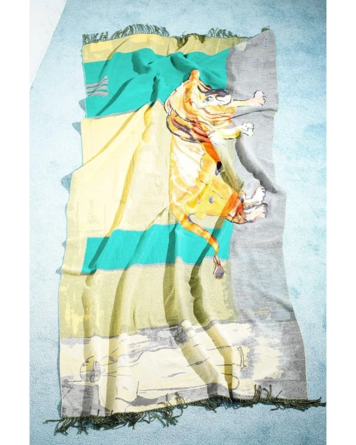 Abstract blanket with tiger graphic in yellow, orange, teal, and grey tones with fringe on both ends, flat lay on light blue backdrop.