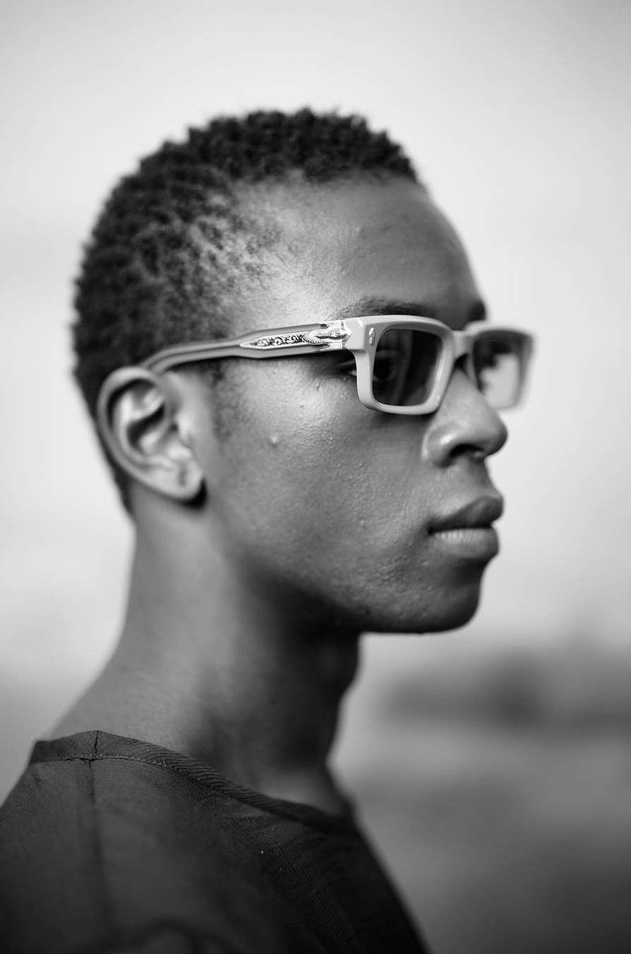 Profile portrait of a black man wearing light-framed glasses and a dark shirt against an outdoor background in Paris