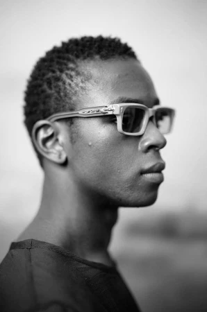 Profile portrait of a black man wearing light-framed glasses and a dark shirt against an outdoor background in Paris