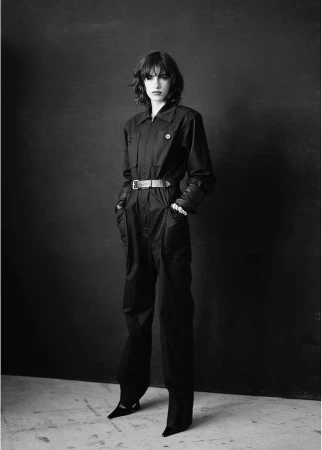 Model in black jumpsuit with a belted waist standing against a dark textured wall in a black and white portrait.