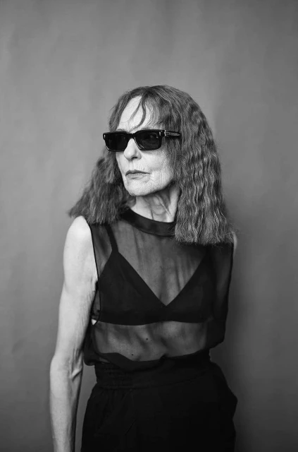 Black-and-white studio portrait of an elderly woman with long hair, wearing sunglasses, a sheer top, and dark pants against a gray backdrop.