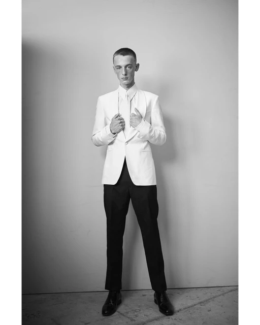 Full-length portrait of a young man in a white blazer and black pants, posing against a neutral background in a black and white photograph.