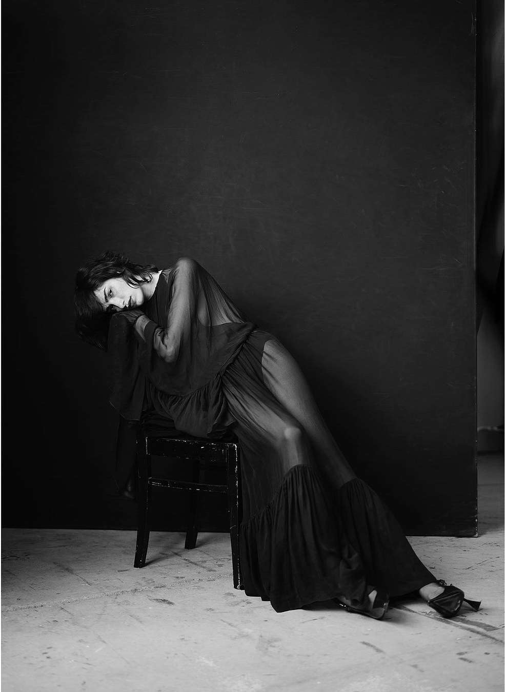 A woman in a sheer black dress reclines on a chair in a black and white fashion shot with low-key lighting against a dark backdrop.
